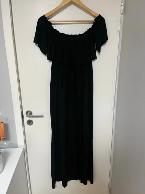 Svart offshoulder långklänning - Svart långklänning med offshoulder-design och volang upptill. Klänningen har en luftig passform och är perfekt för sommarens varma dagar. Tillverkad i ett mjukt och följsamt material som ger en elegant siluett. Ganska så hög slits. Det står ingen storlek men den passar M/L.