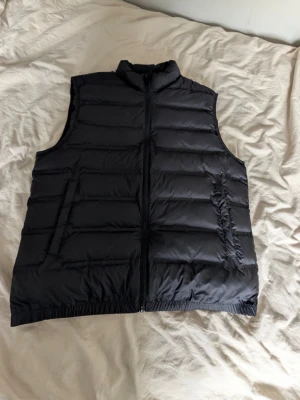 Zara väst - Stilren svart dunväst med quiltad design och hög krage. Västen har dragkedja framtill och två sidofickor. Perfekt att slänga över en hoodie eller tröja för extra värme. Enkel och clean look som passar till det mesta.