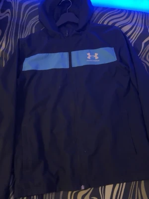 Mörk blå vindjacka och byxor. - Svart vindjacka med blå paneler över bröstet och Under Armour-logga. Jackan har huva, dragkedja framtill och en sportig look. Perfekt för blåsiga dagar och enkel att matcha med det mesta.