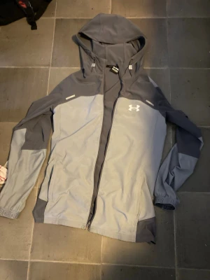 Grå vindjacka från Under Armour - Snygg tvåfärgad grå vindjacka från Under Armour med huva och dragkedja framtill. Jackan har logga på bröstet och stor text på ryggen. Tillverkad i lätt och tåligt material, perfekt för blåsiga dagar. Justerbara muddar och fickor med dragkedja.