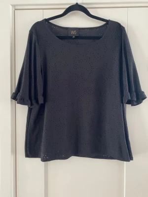 Svart blus med broderade detaljer W5 - Svart blus från W5 med broderade hålmönster och vida trekvartsärmar med volangkant. Blusen har rund halsringning och är tillverkad i ett mjukt bomullstyg som ger en luftig känsla. Perfekt för dig som gillar stilrena men ändå detaljerade plagg. Använd men i fint begagnat skick   Mått Över bröstet: ca 57 cm Längd från axeln och ner: ca 61 cm  Kommer från ett djur och rökfritt hem