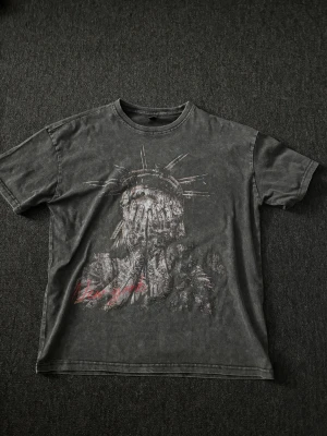 Svart topp med Frihetsgudinnan-tryck - Snygg svart topp i bomull med ett stort tryck av Frihetsgudinnan som täcker ansiktet. T-shirten har en tvättad vintage-look, rund hals och normal passform. Röd textdetalj framtill ger extra edge till stilen.