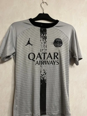 Grå PSG matchtröja Nike M - Snygg grå PSG matchtröja från Nike med svart vertikal rand och Paris Saint-Germain-logga på bröstet. Tröjan har korta ärmar, svart krage och Qatar Airways-tryck på framsidan. Tillverkad i lätt och ventilerande material, perfekt för fotboll eller träning.