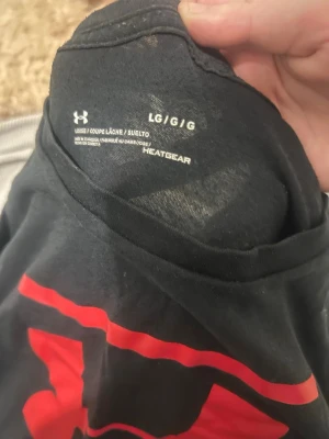 Svart Under Armour t-shirt med logga - Snygg svart t-shirt från Under Armour med stor röd och vit logga på bröstet. Tillverkad i ett lätt och funktionellt material som andas, perfekt för träning eller chill. Modellen är loose fit och har korta ärmar.