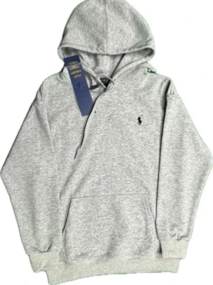 Hoodie Polo Ralph Lauren - Klassisk grå hoodie från Polo Ralph Lauren med broderad logga på bröstet. Hoodien har huva med dragsko, magficka och ribbade muddar vid ärmslut och nederkant. Tillverkad i mjuk bomullsblandning för skön komfort. Perfekt för en avslappnad och stilren look.