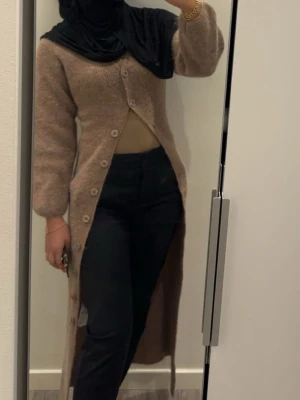 Lång beigebrun kofta med knappar - Säljer en lång, beigebrun kofta med stora knappar framtill. Koftan har en rak passform, långa ärmar och är stickad i ett mjukt material. Passar som klänning eller kofta, framhäver kroppen fint vid användning som klänning. Perfekt att slänga över en outfit för extra värme och stil. Enbart använd i några timmar