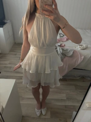 Vit omlottklänning med volanger studentklänning - Säljer en vit kortklänning med halterneck och omlottdesign. Klänningen har flera volanger. Storlek xs. Oanvänd och taggen sitter kvar 