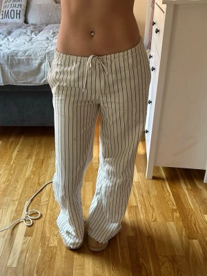 Vita linnebyxor med svarta ränder - Säljer ett par vita linnebyxor med tunna svarta vertikala ränder och dragsko i midjan. Byxorna har en avslappnad passform och raka ben, perfekta för en chill och stilren look. Materialet är luftigt och skönt, perfekt för varma dagar.