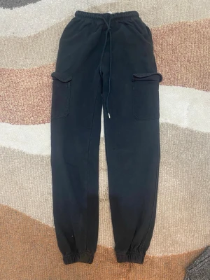Svarta cargomjukisbyxor med fickor - Säljer ett par svarta mjukisbyxor med cargofickor på sidorna och resår i midjan samt vid bensluten. Byxorna har dragsko i midjan för skön passform och är tillverkade i mjukt sweatshirtmaterial. Perfekta för en avslappnad och trendig look.