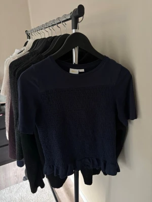 Mörkblå topp med volangkant - Snygg mörkblå topp med korta ärmar och smockad framsida. Toppen har en söt volangkant nedtill och rund halsringning. Perfekt att matcha med både jeans och kjol för en trendig look.