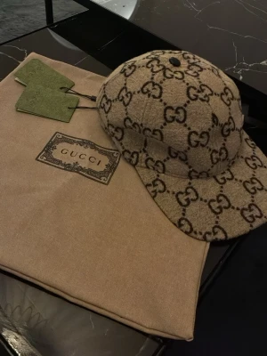 Beige keps från Gucci med GG-mönster - Säljer en beige keps från Gucci med det klassiska GG-monogrammet i brunt över hela kepsen. Tillverkad i Italien och har en böjd skärm. Kepsen kommer med originalpåse och tags, vilket ger en exklusiv känsla.