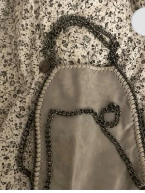 Grå handväska med kedjerem - Snygg grå handväska i mockaimitation med dekorativa kedjedetaljer längs kanterna och som axelrem. Väskan har en mjuk, slät yta och en rundad form. Perfekt för dig som gillar trendiga accessoarer med lite edge.