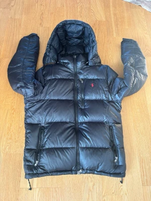 Svart pufferjacka från Polo Ralph Lauren - Snygg svart pufferjacka från Polo Ralph Lauren med röd broderad logga på bröstet. Jackan har huva, två dragkedjeförsedda fickor och en glansig finish. Perfekt för kalla dagar och riktigt varm tack vare det tjocka materialet.