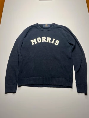 Mörkblå stickad tröja från Morris - Mörkblå stickad tröja från Morris med vit text på bröstet. Tröjan har rund halsringning och ribbstickade muddar vid ärmslut och nederkant. Perfekt för en stilren och avslappnad look.