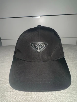 Svart keps från Prada med logga - Svart keps från Prada i klassisk modell med böjd skärm och justerbar rem bak. Kepsen har Pradas ikoniska trekantslogga framtill i metall och tyg, vilket ger en lyxig och trendig känsla. Perfekt accessoar för att lyfta din stil.