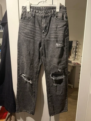 Gråa raka jeans med slitningar - Säljer ett par gråa raka jeans med tydliga slitningar och hål på båda benen. Jeansen har fem fickor, hög midja och klassisk knappgylf. Materialet är denim i bomull och de har en trendig, avslappnad look.