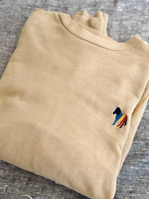 Limiterad Paul Smith sweater - Snygg beige hoodie med en liten broderad häst i flera färger på bröstet. Hoodien har klassisk passform och är tillverkad i mjukt material, perfekt för en chill och stilren look.