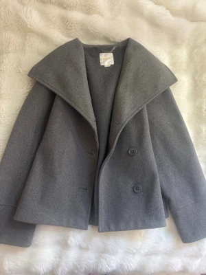 Grå kappa från Gina Tricot - Stilren grå kappa från Gina Tricot med bred krage och dubbelknäppning framtill. Kappan har en rak passform och är tillverkad i ett mjukt, ull-liknande material. Perfekt för att ge en snygg och trendig look under kyliga dagar.