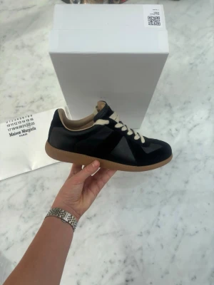 Maison Margiela svarta sneakers - Maison Margiela sneakers i svart läder och mocka med beige detaljer och vita snören. Klassisk låg modell med gummisula i naturfärg. Snygg och stilren design som passar till det mesta.
