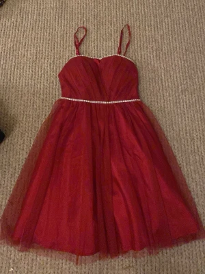Röd kort klänning  - Snygg röd kort klänning med tunna axelband och markerad midja med strassdetalj. Klänningen har ett lätt utställt kjolparti i tyll och en v-ringad överdel. Perfekt för dig som vill sticka ut med en elegant look.