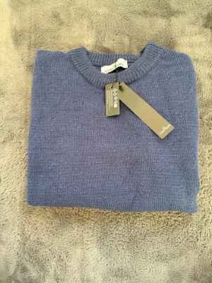 Blå stickad tröja från Stone Island - Snygg blå stickad tröja från Stone Island med rund halsringning och ribbade muddar. Tröjan har en klassisk, enkel design och är tillverkad i mjukt material som passar perfekt till kyligare dagar. Den ser urtvättad ut så kontakta gärna vid efter frågan om fler bilder 
