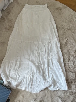 Vit långkjol med resår i midjan - Säljer en vit långkjol med smockad resår i midjan. Kjolen är i ett lätt, vågigt tyg som ger en luftig känsla och har en enkel, stilren design. Perfekt för varma dagar och enkel att matcha med olika toppar. Funkar med högmidja men bäst lågmidjad! Så ledsen att jag behöver sälja den men passar tyvärr inte mig längre❤️storlek 170 men passar perfekt på mig som är ca 164,5