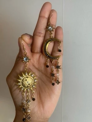 Guldiga örhängen med sol och måne nickel fri - Unika örhängen i guldton med sol- och månform. Dekorerade med små stjärnor, svarta och klara pärlor samt detaljerade kedjor. Perfekt för dig som gillar statement-smycken med magisk vibe och boho-känsla.