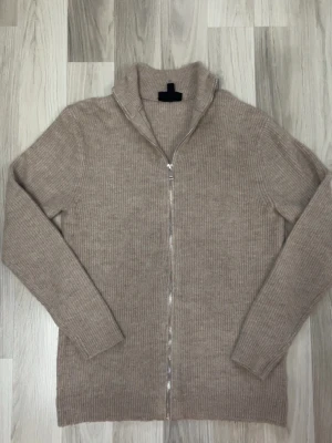  Ziptröja - Beige ribbstickad tröja med dragkedja och hög krage. Tröjan har lång ärm och är tillverkad i ett mjukt, värmande material. Perfekt för lager-på-lager och enkel att matcha med jeans eller chinos.