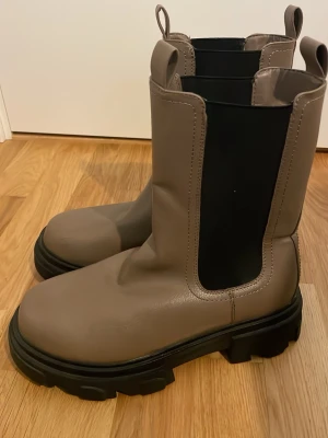 Beige chunky stövletter - Säljer ett par beige chunky stövletter med bred resår i svart på sidorna och grov sula i svart. Stövletterna har dragflikar både fram och bak för enkel påtagning. Tillverkade i ett slätt, matt material som ger en modern look.