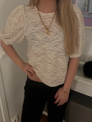 Vit blus med puffärm och struktur - Trendig vit blus med puffärmar och snyggt strukturerat mönster. Blusen har rund halsringning och är tillverkad i ett lätt, genombrutet material som ger en cool look. Perfekt att styla med smycken och svarta byxor.