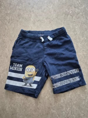 Shorts - Mörkblå shorts med tryck av en Minion och texten 'Team Minion' på vänster ben. Shortsen har vita och blå ränder, samt snörning i midjan för skön passform. Perfekta för sommardagar och chill häng.