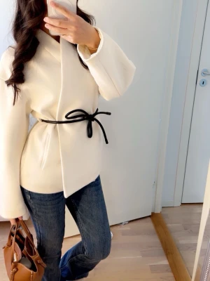 Vit kappa från Kappahl - Stilren krämvit kappa med minimalistisk design och svart knytband i midjan. Kappan har en rak passform, vida ärmar och är tillverkad i ett mjukt tyg som ger en lyxig känsla. Perfekt för dig som gillar enkel men elegant stil. Kommer i väldigt bra skick