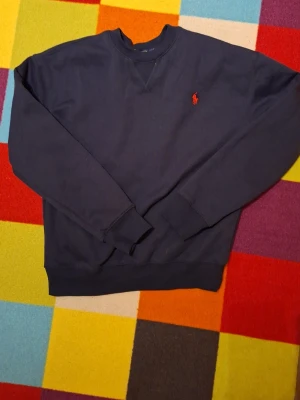 Marinblå sweatshirt Polo Ralph Lauren - Klassisk marinblå sweatshirt från Polo Ralph Lauren med rund halsringning och röd broderad logga på bröstet. Tröjan har ribbade muddar vid ärmslut och nederkant, perfekt för en avslappnad och stilren look. Väldigt bra skick.