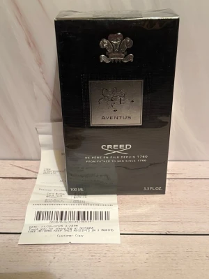 Creed Aventus Eau de Parfum 100ml - Creed Aventus Eau de Parfum är en exklusiv herrdoft i en elegant svart och silver kartong. Flaskan rymmer 100 ml och har en lyxig design med silverdetaljer och klassisk Creed-logga. Doften är känd för sina fruktiga och träiga noter och passar dig som vill sticka ut.