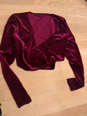 Vinröd omlottopp i sammet från NLY One - Säljer en vinröd omlottopp i mjuk sammet från NLY One. Toppen har lång ärm, v-ringning och croppad passform som ger en trendig look. Perfekt för dig som vill sticka ut med en lyxig känsla och snygg färg.