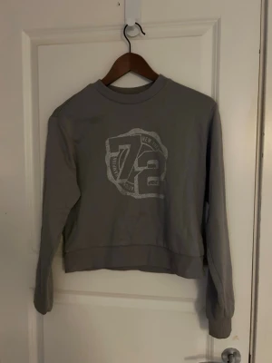 Grå sweatshirt  - Säljer en grå sweatshirt från H&M Divided i storlek XXS. Tröjan har ett stort tryck med siffran 72 och texten 'New York' framtill. Rund hals, ribbade muddar vid ärmslut och nederkant. Perfekt för en chill och avslappnad stil.