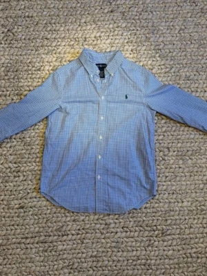 Blårutig skjorta från Ralph Lauren - Klassisk blårutig skjorta från Ralph Lauren med button-down krage och grönt broderat logotyp på bröstet. Skjortan har lång ärm, knappar framtill och är tillverkad i mjuk bomull. Perfekt för en stilren look. Står L men på grund av barnstorlek så passar den som en xs i herrstorlek. 