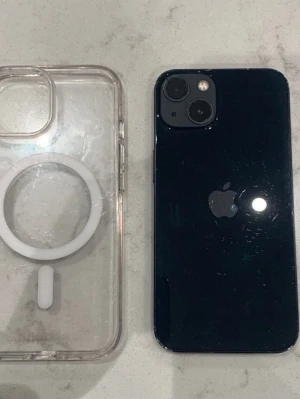 Svart iPhone med MagSafe-skal - Säljer en svart iPhone med stilren design och rundade kanter. Baksidan är i glas och har dubbla kameror. Medföljer ett genomskinligt MagSafe-kompatibelt skal i plast med vit ring för trådlös laddning. Perfekt för dig som gillar Apple och vill ha en snygg mobil.