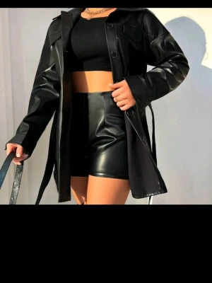 Svart lång skinnjacka med bälte - Snygg svart skinnjacka i längre modell med bälte i midjan. Jackan har klassisk krage, knäppning framtill. Perfekt för en trendig och edgy look. Passar till både jeans och kjol för en cool outfit.