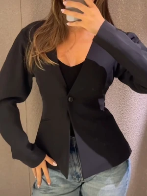 Svart kavaj från Gina Tricot - Stilren svart kavaj från Bik Bok med djup V-ringning och enkel knäppning framtill. Kavajen har raka, långa ärmar och två diskreta fickor. Perfekt att matcha med jeans eller kjol för en trendig look.