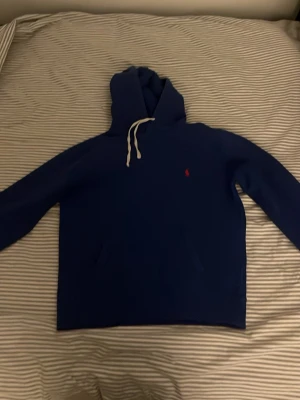 Blå hoodie från Polo Ralph Lauren - Mörkblå hoodie från Polo Ralph Lauren med vit dragsko i huvan och den klassiska röda loggan broderad på bröstet. Hoodien har känguruficka framtill och är gjord i mjukt bomullsmaterial. Perfekt för en chill och stilren look.