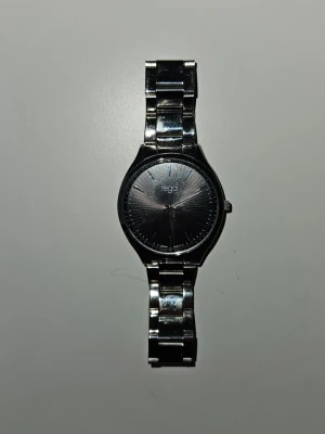 Silverfärgad klocka från Regal - Stilren klocka från Regal med silverfärgat metallarmband och svart urtavla. Klockan har en klassisk rund form och minimalistisk design, perfekt för dig som gillar en clean look. Metallänkarna ger en snygg och modern känsla.