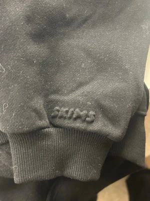 Svart hoodie från Skims - Svart hoodie från Skims med diskret broderad logga på ärmen. Klassisk modell med ribbade muddar och mjukt material som känns skönt mot huden. Perfekt för en avslappnad och stilren look.