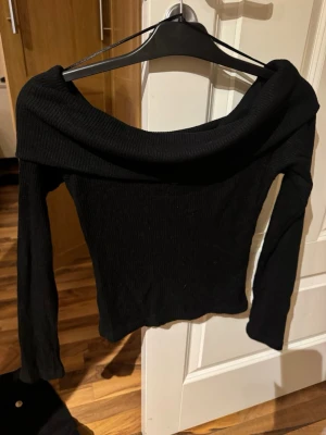 Svart off shoulder topp - Trendig svart off shoulder topp med långa ärmar och ribbad struktur. Sitter tajt och har en bred, vikt krage som ger en snygg och stilren look. Perfekt att matcha med jeans eller kjol för en cool outfit.