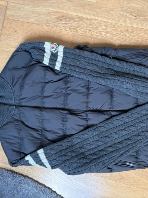 Grå cardiganjacka från Moncler - Snygg grå cardiganjacka från Moncler med stickade ärmar och rygg i kabelstickat mönster. Framsidan är quiltad och vadderad för extra värme. Ärmarna har vita ränder och Moncler-logga på axeln. Dragkedja framtill och hög krage.