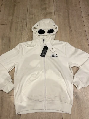 Vit hoodie med goggles från C.P. Company - Säljer en vit hoodie från C.P. Company med deras ikoniska goggles i huvan. Tröjan har dragkedja framtill, två stora fickor och logotyp på bröstet. Materialet är mjukt och bekvämt, perfekt för en cool och avslappnad look.