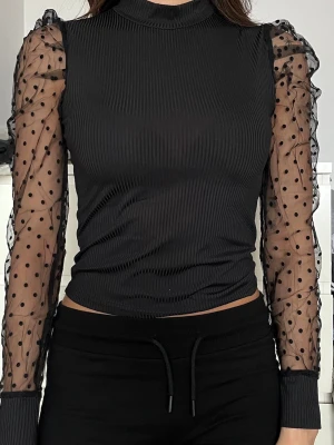Svart topp med prickiga meshärmar - Trendig svart topp med ribbad kropp och transparenta meshärmar med svarta prickar. Toppen har hög hals och figurnära passform, perfekt för en snygg och modern look. Ärmarna är långa och ger en cool kontrast till den enfärgade kroppen.