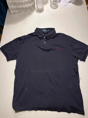 Svart pikétröja från Polo Ralph Lauren - Klassisk svart pikétröja från Polo Ralph Lauren med korta ärmar och krage. Tröjan har knappar framtill och den ikoniska röda logotypen broderad på bröstet. Tillverkad i mjuk bomull som är skön att bära.
