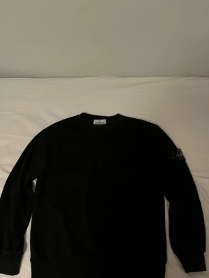 Svart sweatshirt från Stone Island - Säljer en svart sweatshirt från Stone Island med den klassiska kompasspatchen på vänster ärm. Tröjan har rund halsringning och långa ärmar. Tillverkad i mjukt bomullsmaterial som känns skönt mot huden. Perfekt för dig som gillar streetwear och vill ha en clean look.