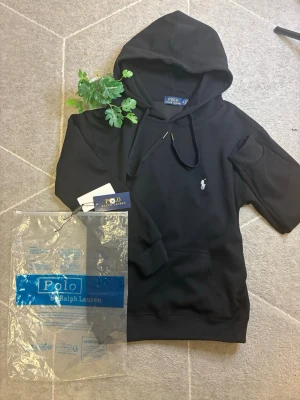 Svart hoodie från Polo Ralph Lauren - Snygg svart hoodie från Polo Ralph Lauren med klassisk huva och dragsko. Ikoniska vita logon broderad på bröstet. Tillverkad i mjukt material med ribbade muddar vid ärmar och nederkant. Perfekt för en avslappnad och stilren look.